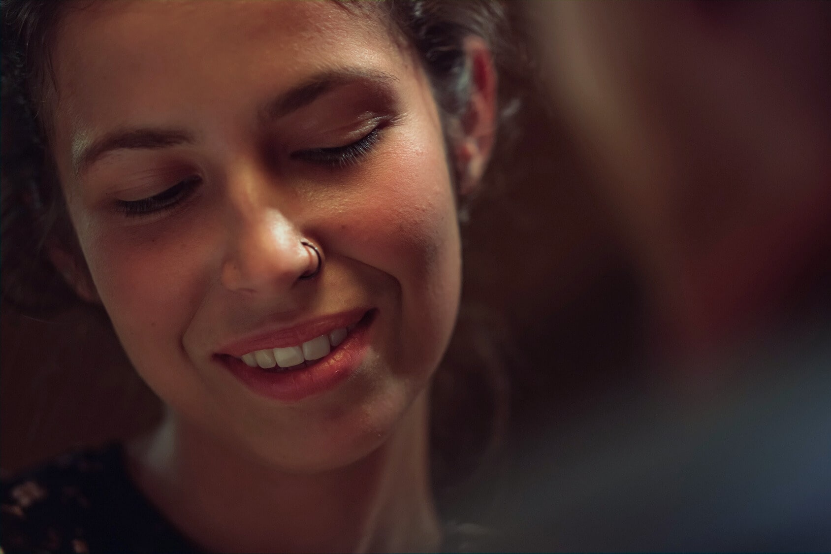 A person with curly hair and a nose ring is smiling gently, looking downward. Soft lighting creates a warm ambiance, highlighting their relaxed and contented expression.