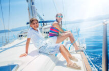 Two smiling people, a boy in a white shirt and shorts and a woman in a striped tank top and shorts, sitting on the deck of a sailboat. They're wearing sunglasses and enjoying a sunny day at sea with a clear blue sky and calm water.