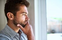 A man with short brown hair and a beard stands indoors, looking thoughtfully out of a window. He is wearing a light grey button-up shirt, and his hand rests on his chin in a contemplative pose.