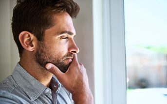 A man with short brown hair and a beard stands indoors, looking thoughtfully out of a window. He is wearing a light grey button-up shirt, and his hand rests on his chin in a contemplative pose.