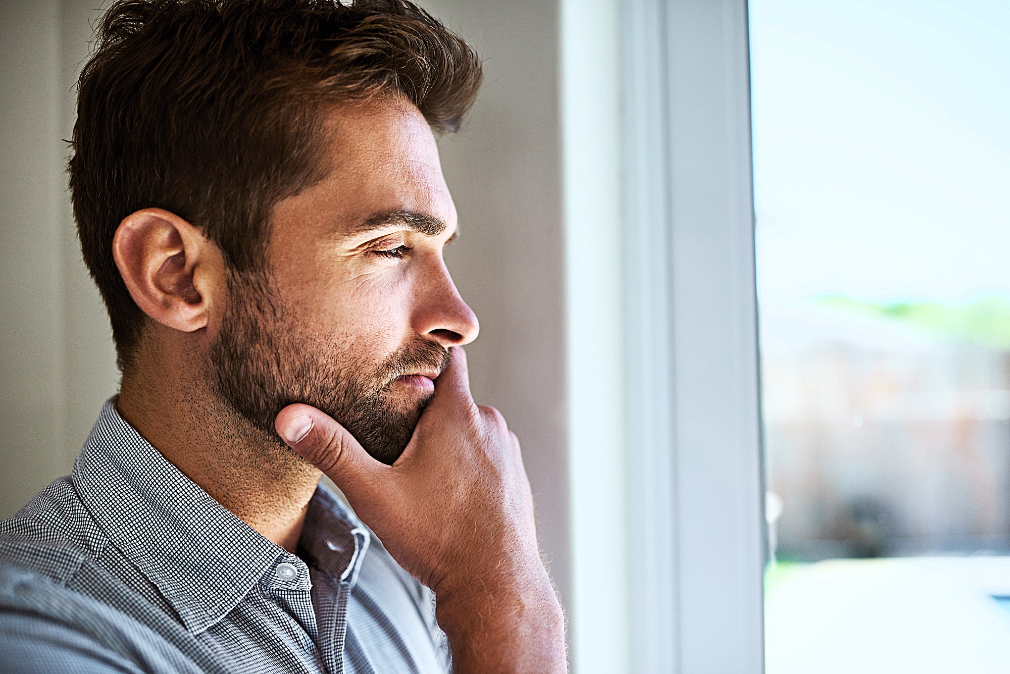 A man with short brown hair and a beard stands indoors, looking thoughtfully out of a window. He is wearing a light grey button-up shirt, and his hand rests on his chin in a contemplative pose.