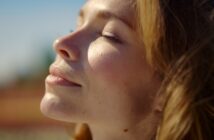 Close-up of a woman with closed eyes basking in sunlight, with a serene expression on her face. Her light brown hair gently frames her face against a blurred outdoor background.