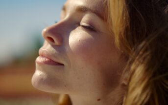 Close-up of a woman with closed eyes basking in sunlight, with a serene expression on her face. Her light brown hair gently frames her face against a blurred outdoor background.