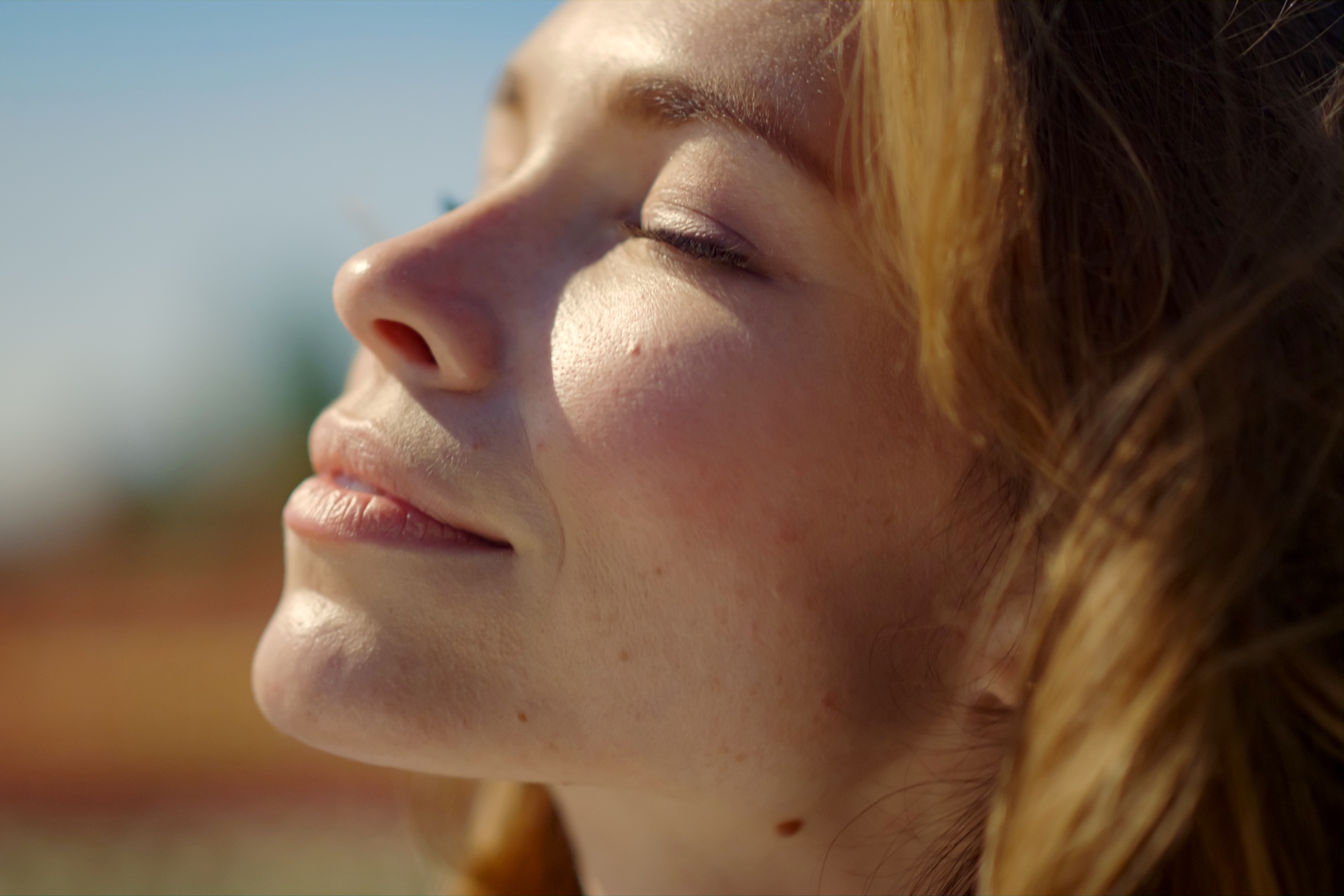 Close-up of a woman with closed eyes basking in sunlight, with a serene expression on her face. Her light brown hair gently frames her face against a blurred outdoor background.