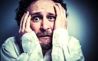 A man with curly hair and a beard holds his head with both hands, his eyes wide open and mouth slightly agape, expressing a look of panic or anxiety. He is wearing a white dress shirt.