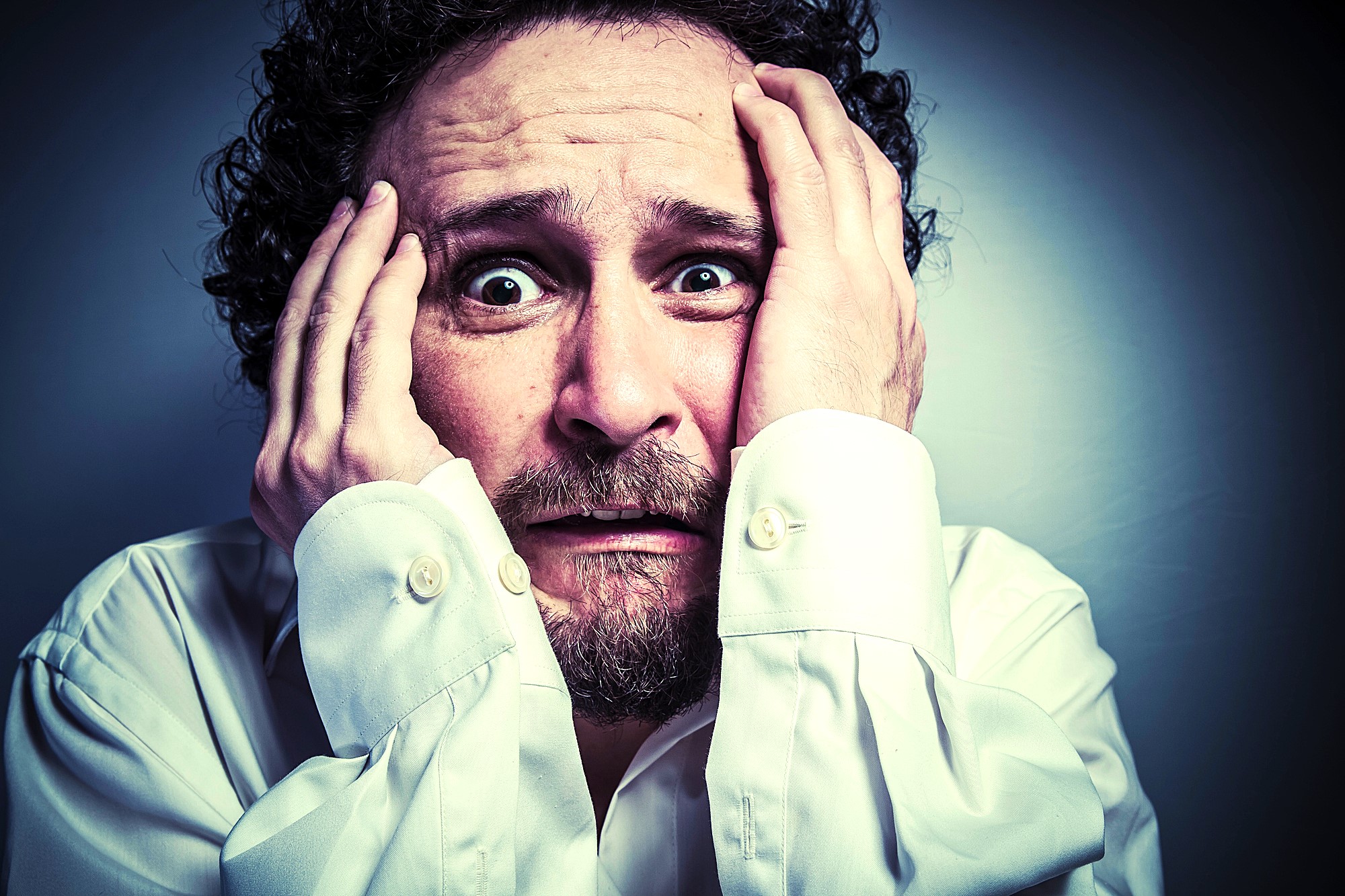 A man with curly hair and a beard holds his head with both hands, his eyes wide open and mouth slightly agape, expressing a look of panic or anxiety. He is wearing a white dress shirt.