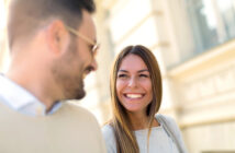 A man and a woman are smiling and looking at each other while standing outdoors. The woman has long hair and is wearing a light-colored sweater. The man is bearded, wearing glasses, and dressed in a light-colored top. The background is a blurred building facade.