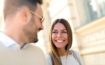 A man and a woman are smiling and looking at each other while standing outdoors. The woman has long hair and is wearing a light-colored sweater. The man is bearded, wearing glasses, and dressed in a light-colored top. The background is a blurred building facade.