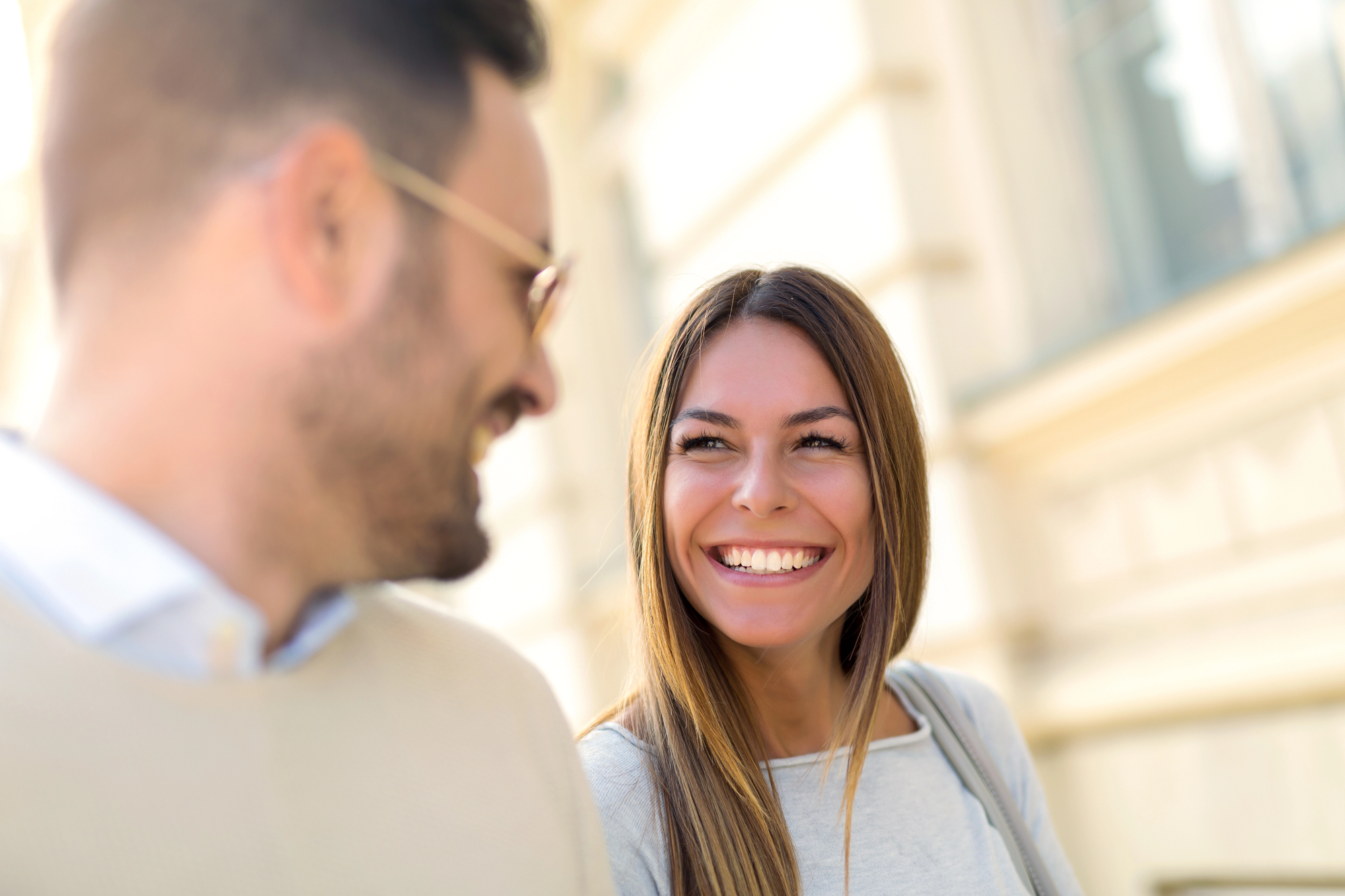 A man and a woman are smiling and looking at each other while standing outdoors. The woman has long hair and is wearing a light-colored sweater. The man is bearded, wearing glasses, and dressed in a light-colored top. The background is a blurred building facade.