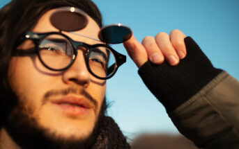 A person with long hair wears round, clear glasses with additional tinted lenses flipped up. They are gazing upward, holding the lenses with a gloved hand. The sky is clear and blue, suggesting sunny weather.