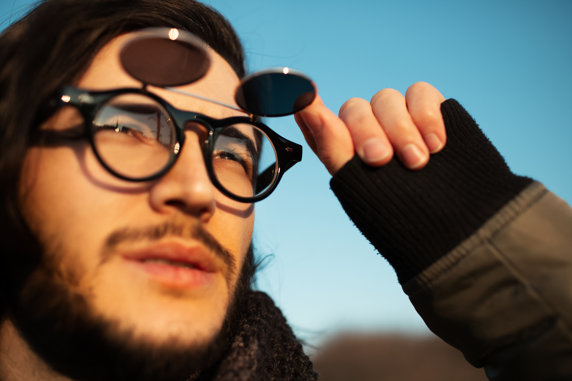 A person with long hair wears round, clear glasses with additional tinted lenses flipped up. They are gazing upward, holding the lenses with a gloved hand. The sky is clear and blue, suggesting sunny weather.