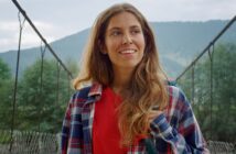 A woman with long hair smiles while standing on a suspension bridge. She is wearing a red shirt and a plaid jacket. The background features a mountainous landscape with greenery and a cloudy sky.