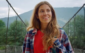 A woman with long hair smiles while standing on a suspension bridge. She is wearing a red shirt and a plaid jacket. The background features a mountainous landscape with greenery and a cloudy sky.