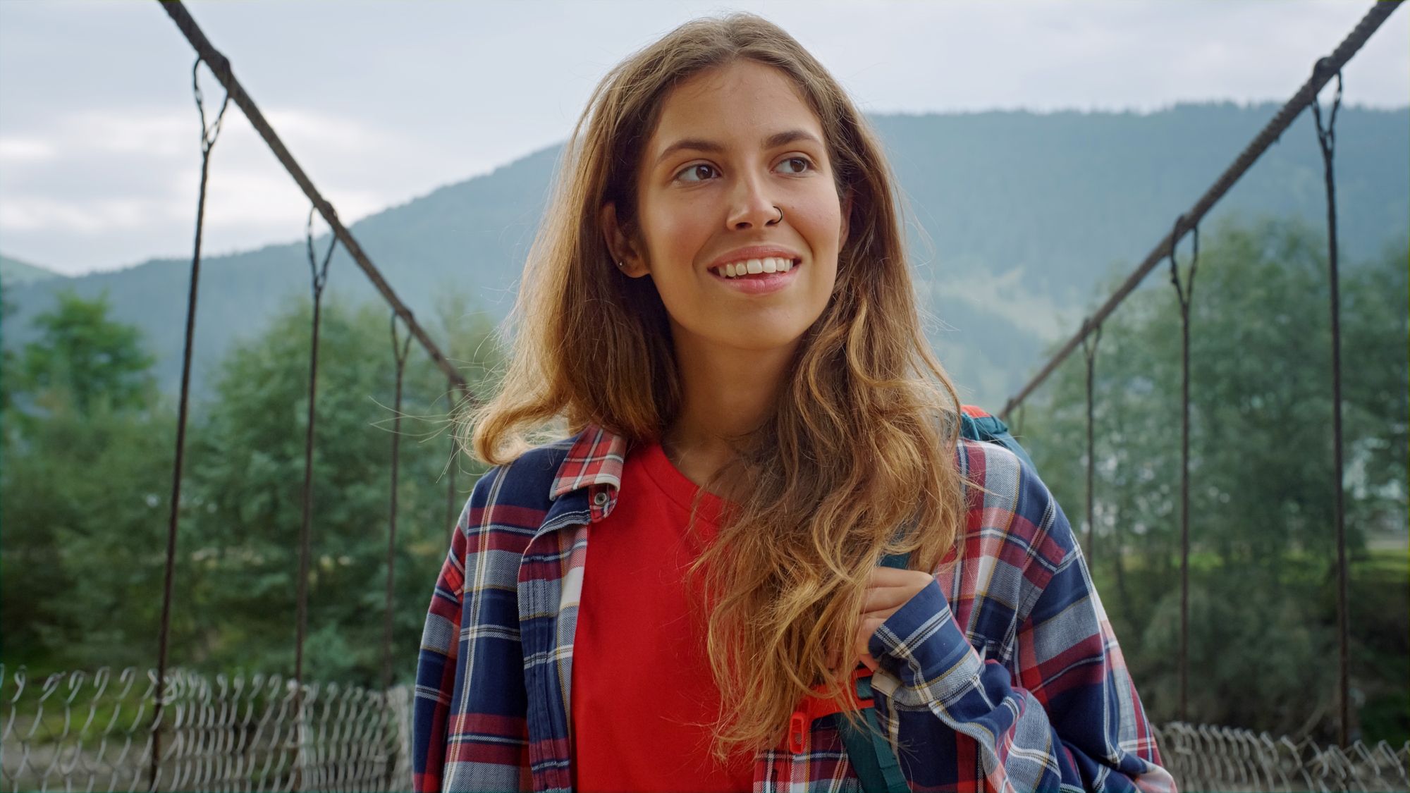 A woman with long hair smiles while standing on a suspension bridge. She is wearing a red shirt and a plaid jacket. The background features a mountainous landscape with greenery and a cloudy sky.