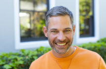 A man with short gray hair and a beard is smiling at the camera. He is wearing an orange sweater and standing outdoors in front of a building with large windows and greenery.