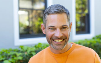 A man with short gray hair and a beard is smiling at the camera. He is wearing an orange sweater and standing outdoors in front of a building with large windows and greenery.