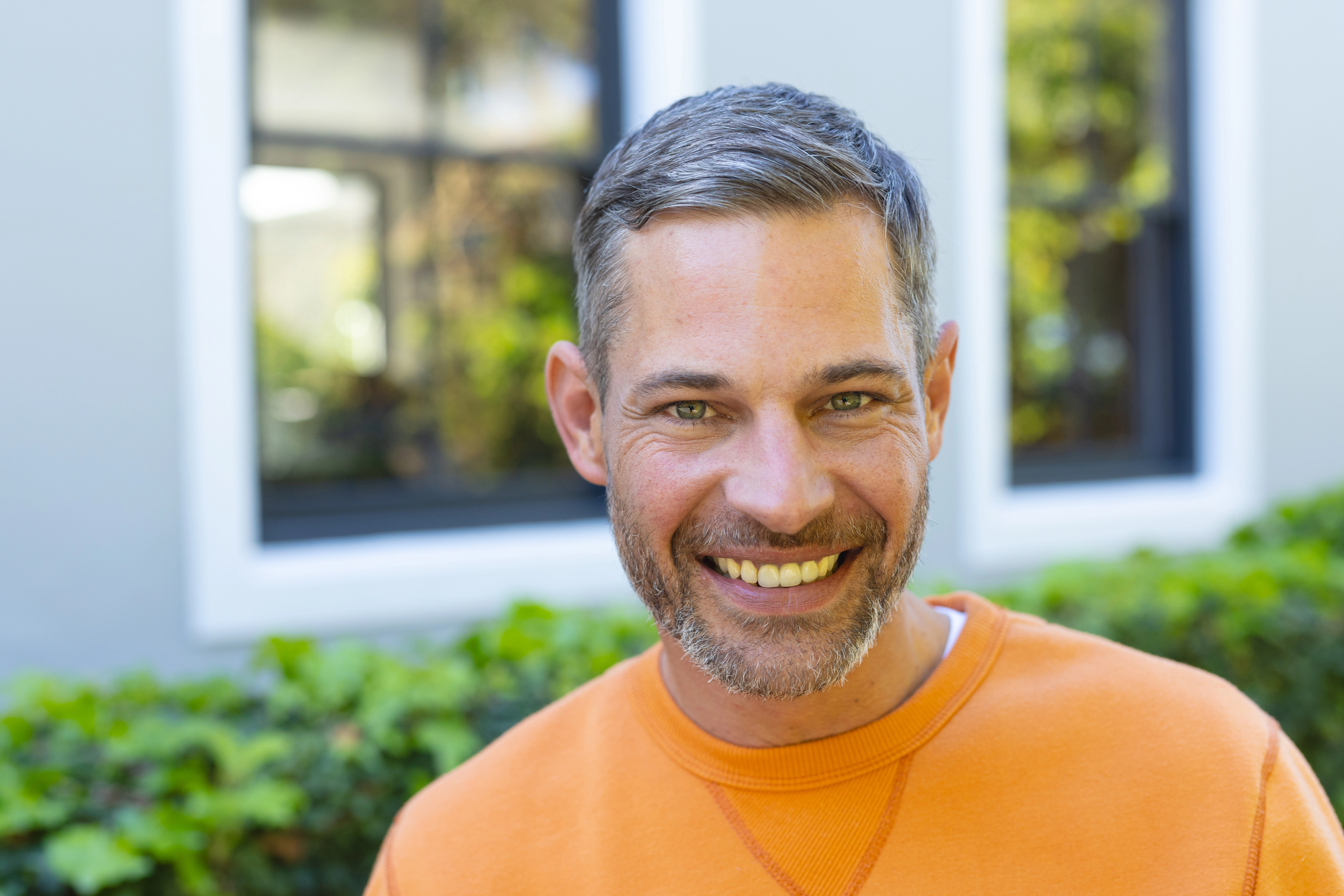 A man with short gray hair and a beard is smiling at the camera. He is wearing an orange sweater and standing outdoors in front of a building with large windows and greenery.