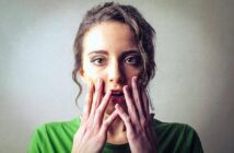 A woman with long hair in a green shirt looks surprised, holding her hands close to her mouth. The background is a neutral shade, highlighting her expression.