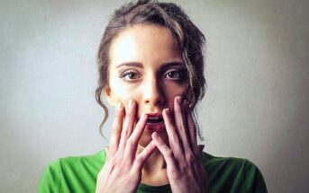 A woman with long hair in a green shirt looks surprised, holding her hands close to her mouth. The background is a neutral shade, highlighting her expression.