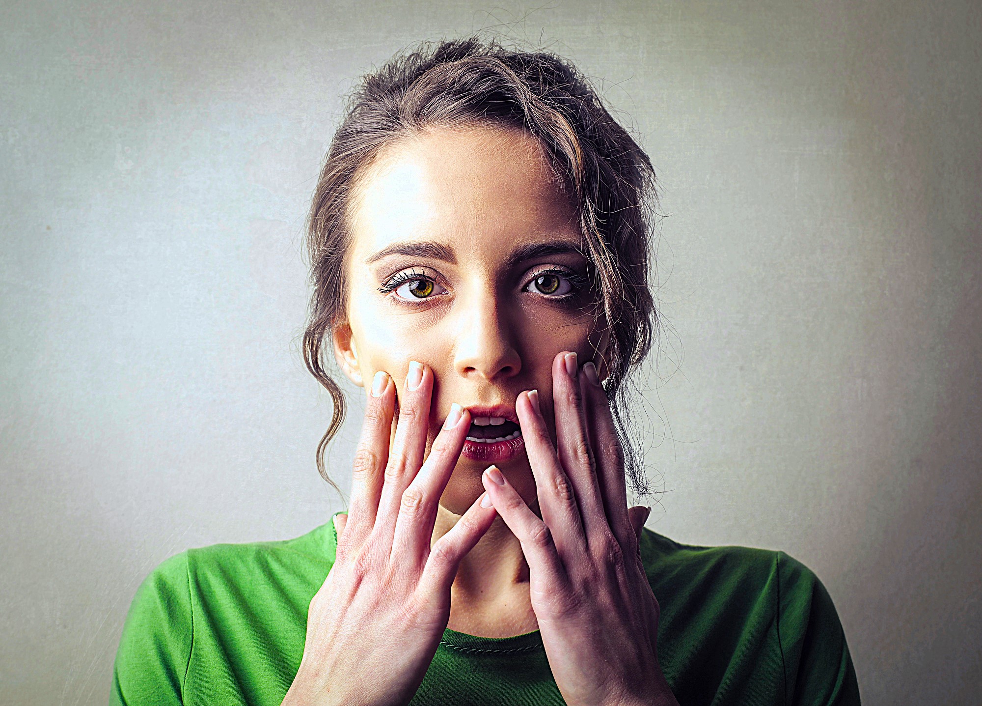 A woman with long hair in a green shirt looks surprised, holding her hands close to her mouth. The background is a neutral shade, highlighting her expression.