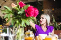 A woman with blonde hair in a purple top is raising her arm and speaking animatedly in a restaurant. She is surrounded by vibrant pink flowers, plants, and glass teapots on the table. The setting has a warm, rustic ambiance.