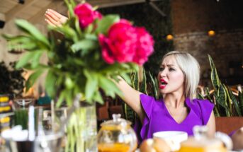 A woman with blonde hair in a purple top is raising her arm and speaking animatedly in a restaurant. She is surrounded by vibrant pink flowers, plants, and glass teapots on the table. The setting has a warm, rustic ambiance.