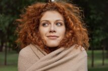 A person with curly red hair wrapped in a beige blanket stands outdoors, looking to the side with a slight smile. Green trees are visible in the blurred background.