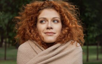 A person with curly red hair wrapped in a beige blanket stands outdoors, looking to the side with a slight smile. Green trees are visible in the blurred background.