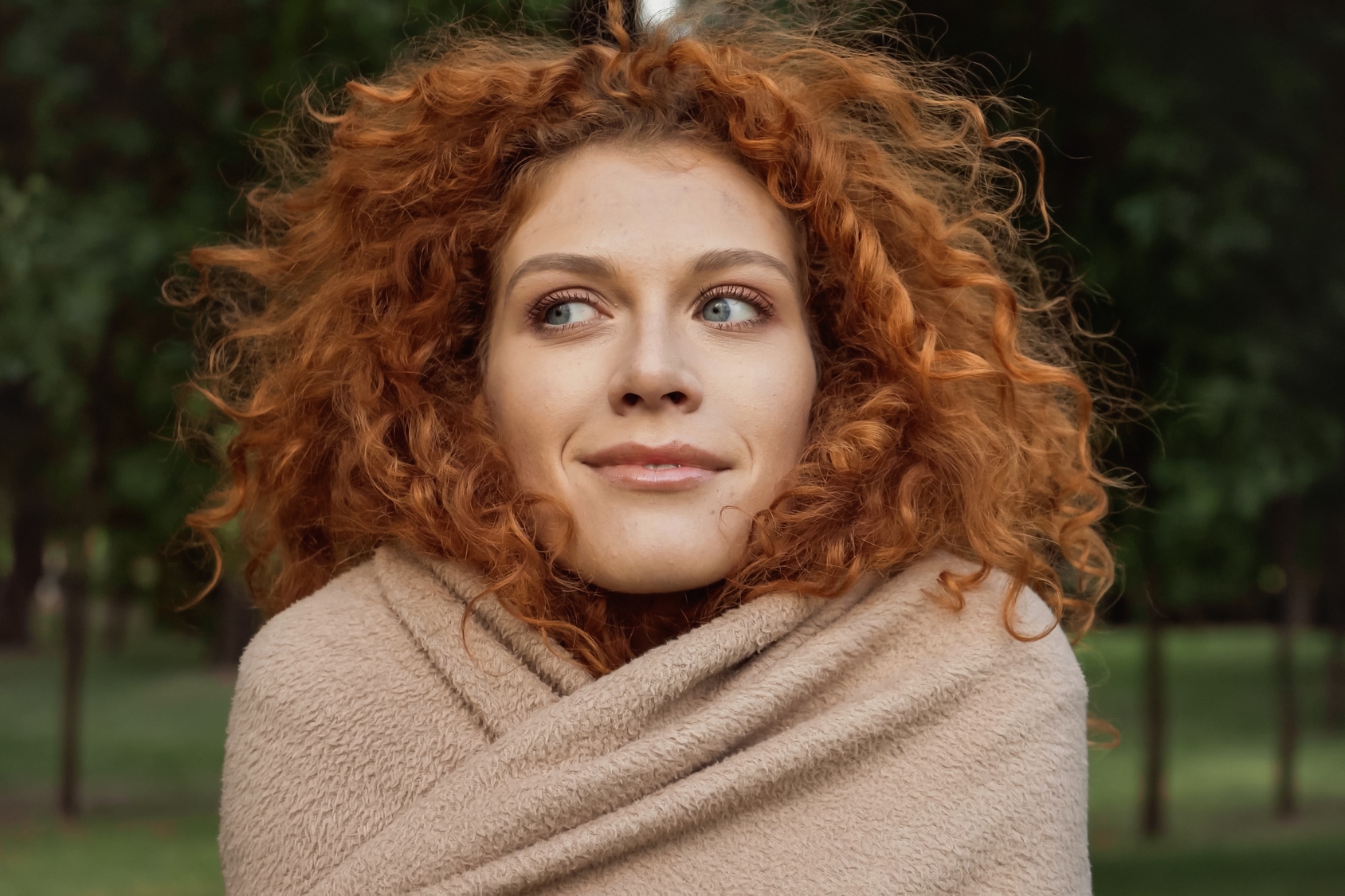 A person with curly red hair wrapped in a beige blanket stands outdoors, looking to the side with a slight smile. Green trees are visible in the blurred background.