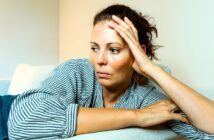 A woman with tied-back hair, wearing a striped shirt, leans on a light-colored couch with her head resting on her hand, looking contemplative and to the side. The wall behind her is plain and light-colored.