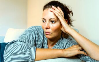 A woman with tied-back hair, wearing a striped shirt, leans on a light-colored couch with her head resting on her hand, looking contemplative and to the side. The wall behind her is plain and light-colored.