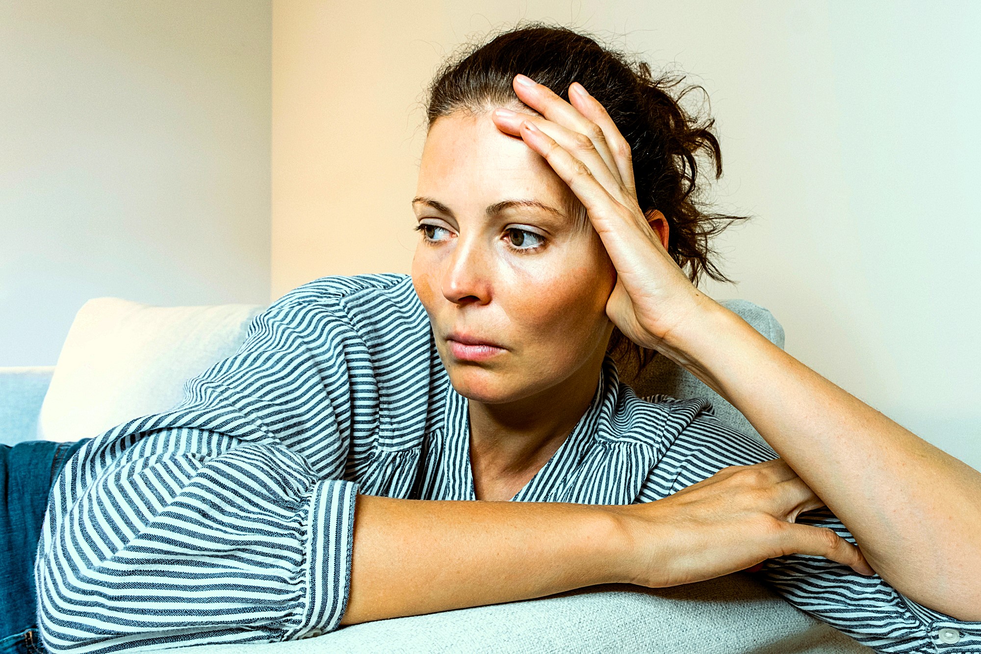 A woman with tied-back hair, wearing a striped shirt, leans on a light-colored couch with her head resting on her hand, looking contemplative and to the side. The wall behind her is plain and light-colored.