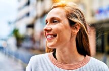 A woman with long hair smiles and looks to the side while standing outdoors. She is wearing a light sweater with a pink collar. The background is a blurred urban setting.