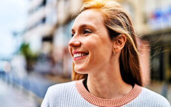 A woman with long hair smiles and looks to the side while standing outdoors. She is wearing a light sweater with a pink collar. The background is a blurred urban setting.