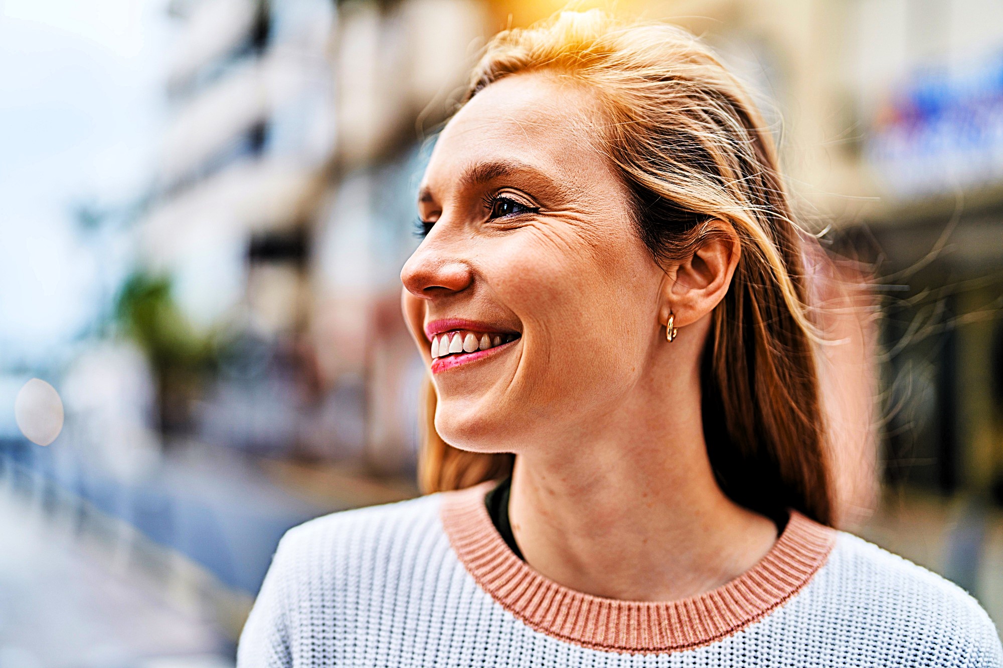 A woman with long hair smiles and looks to the side while standing outdoors. She is wearing a light sweater with a pink collar. The background is a blurred urban setting.