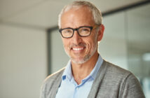 A smiling older man with gray hair and glasses is wearing a blue shirt and gray cardigan. He stands in a modern office with a glass wall in the background.