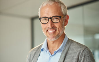 A smiling older man with gray hair and glasses is wearing a blue shirt and gray cardigan. He stands in a modern office with a glass wall in the background.