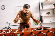 A bearded man passionately playing foosball, wearing a brown sweater and standing near a table with a bicycle and shelves in the background.