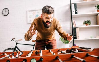 A bearded man passionately playing foosball, wearing a brown sweater and standing near a table with a bicycle and shelves in the background.