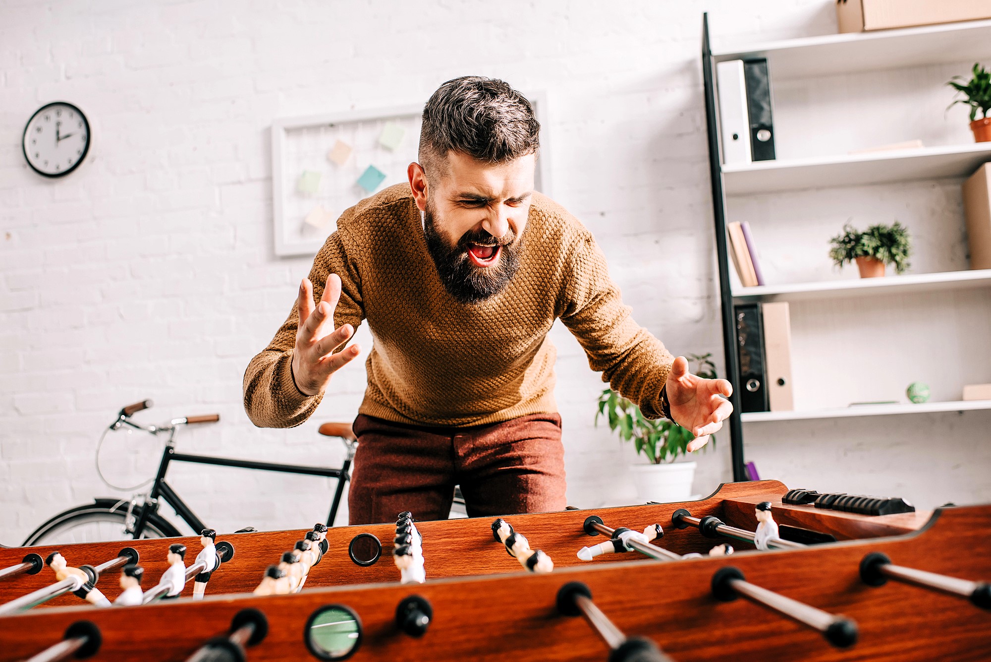 A bearded man passionately playing foosball, wearing a brown sweater and standing near a table with a bicycle and shelves in the background.