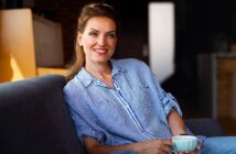 A woman with brown hair, wearing a blue striped shirt, sits on a couch holding a white mug. She is smiling and relaxed, in a warmly lit interior space with a blurred background.