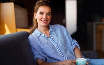 A woman with brown hair, wearing a blue striped shirt, sits on a couch holding a white mug. She is smiling and relaxed, in a warmly lit interior space with a blurred background.