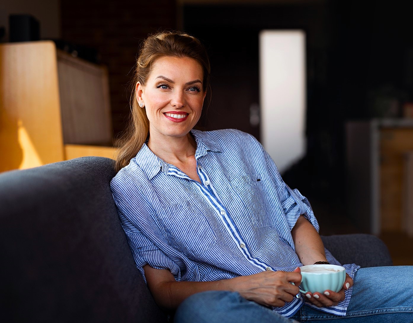 A woman with brown hair, wearing a blue striped shirt, sits on a couch holding a white mug. She is smiling and relaxed, in a warmly lit interior space with a blurred background.