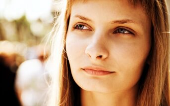 A young woman with long blonde hair gazes thoughtfully into the distance. The background is blurred with warm, natural lighting, creating a serene and introspective atmosphere.