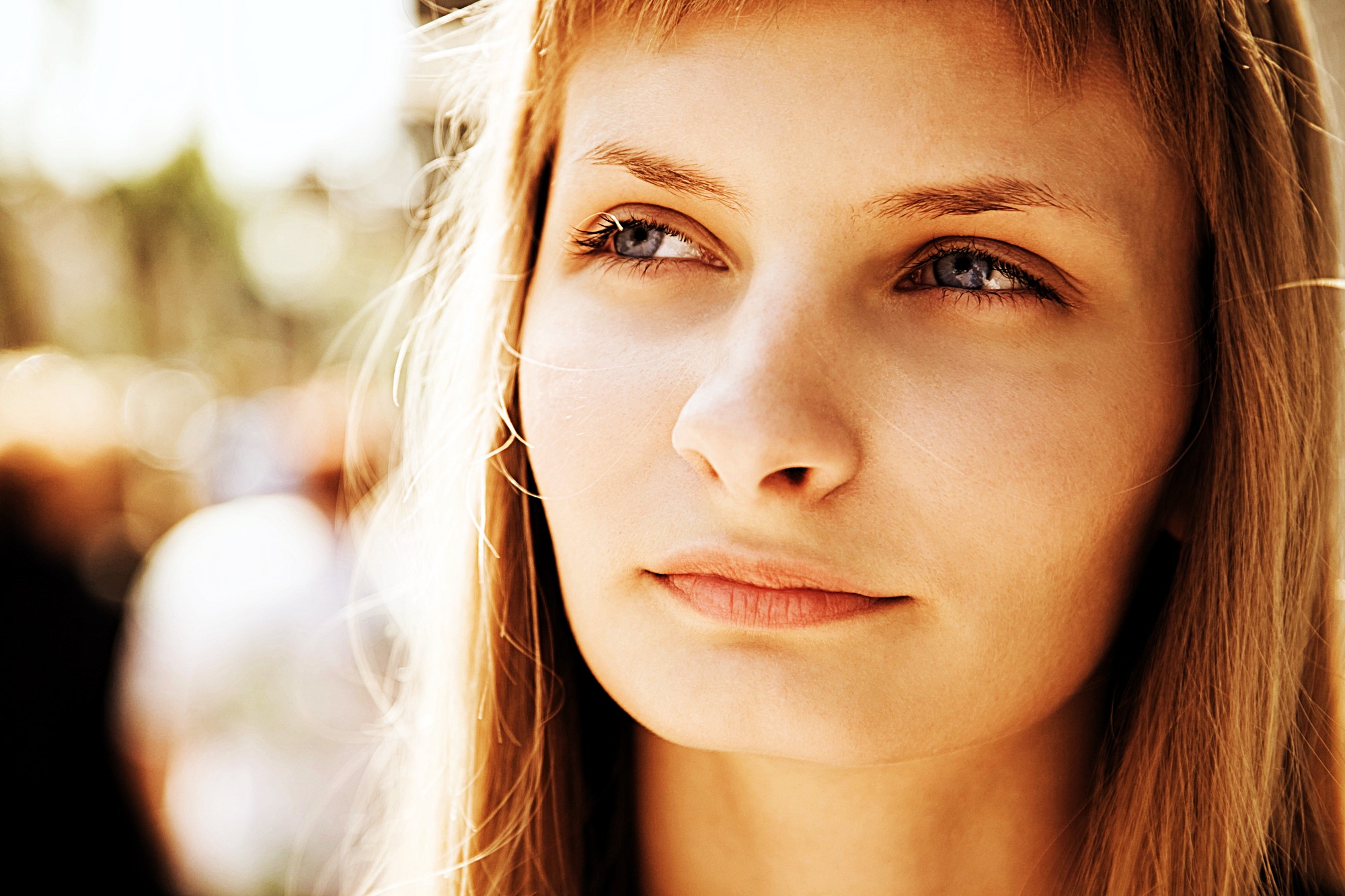 A young woman with long blonde hair gazes thoughtfully into the distance. The background is blurred with warm, natural lighting, creating a serene and introspective atmosphere.