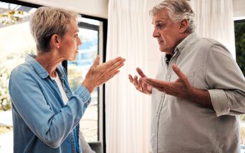 An older man and woman are having a heated argument in a brightly lit room. The woman, with short blonde hair, gestures with open hands. The man, with gray hair, looks frustrated, raising his palms upward. They stand near large windows with curtains.