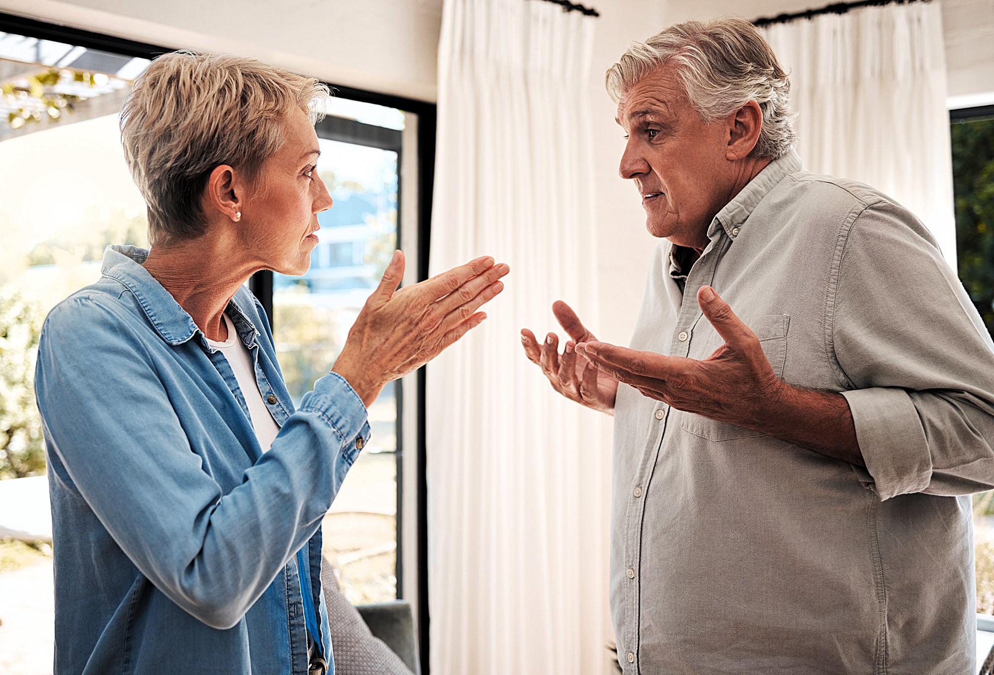 An older man and woman are having a heated argument in a brightly lit room. The woman, with short blonde hair, gestures with open hands. The man, with gray hair, looks frustrated, raising his palms upward. They stand near large windows with curtains.