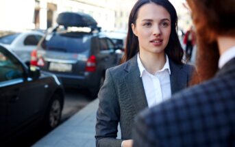 A woman in a gray suit and white shirt stands on a city sidewalk, engaged in conversation. Cars are parked along the street in the background, and she appears focused and attentive to the person she's speaking with.