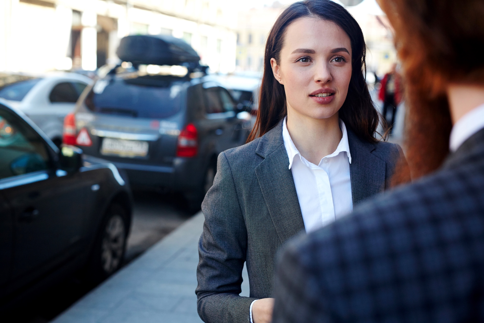 A woman in a gray suit and white shirt stands on a city sidewalk, engaged in conversation. Cars are parked along the street in the background, and she appears focused and attentive to the person she's speaking with.