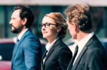 Three professionals, two men and one woman, walk outdoors wearing formal suits. The woman in the center has glasses and a confident expression. They appear engaged in conversation against a blurred urban background.